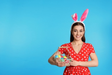 Beautiful woman in bunny ears headband holding basket with Easter eggs on color background, space for text