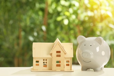 Photo of Piggy bank and house model on wooden table outdoors, space for text. Saving money concept