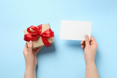 Photo of Woman holding gift box and blank card on light blue background, top view