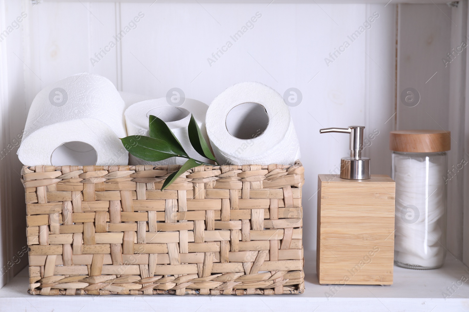 Photo of Toilet paper rolls in wicker basket, green leaves, cotton pads and dispenser on white shelf
