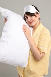 Woman in pyjama and sleep mask holding pillow on grey background