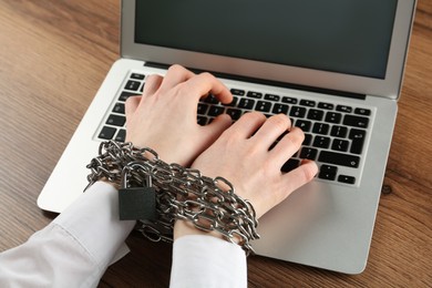 Photo of Woman with chained hands typing on laptop at wooden table, closeup. Internet addiction