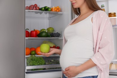 Young pregnant woman with apple near fridge at home, closeup. Healthy eating
