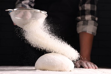 Photo of Man sprinkling flour over dough at wooden table on dark background, closeup