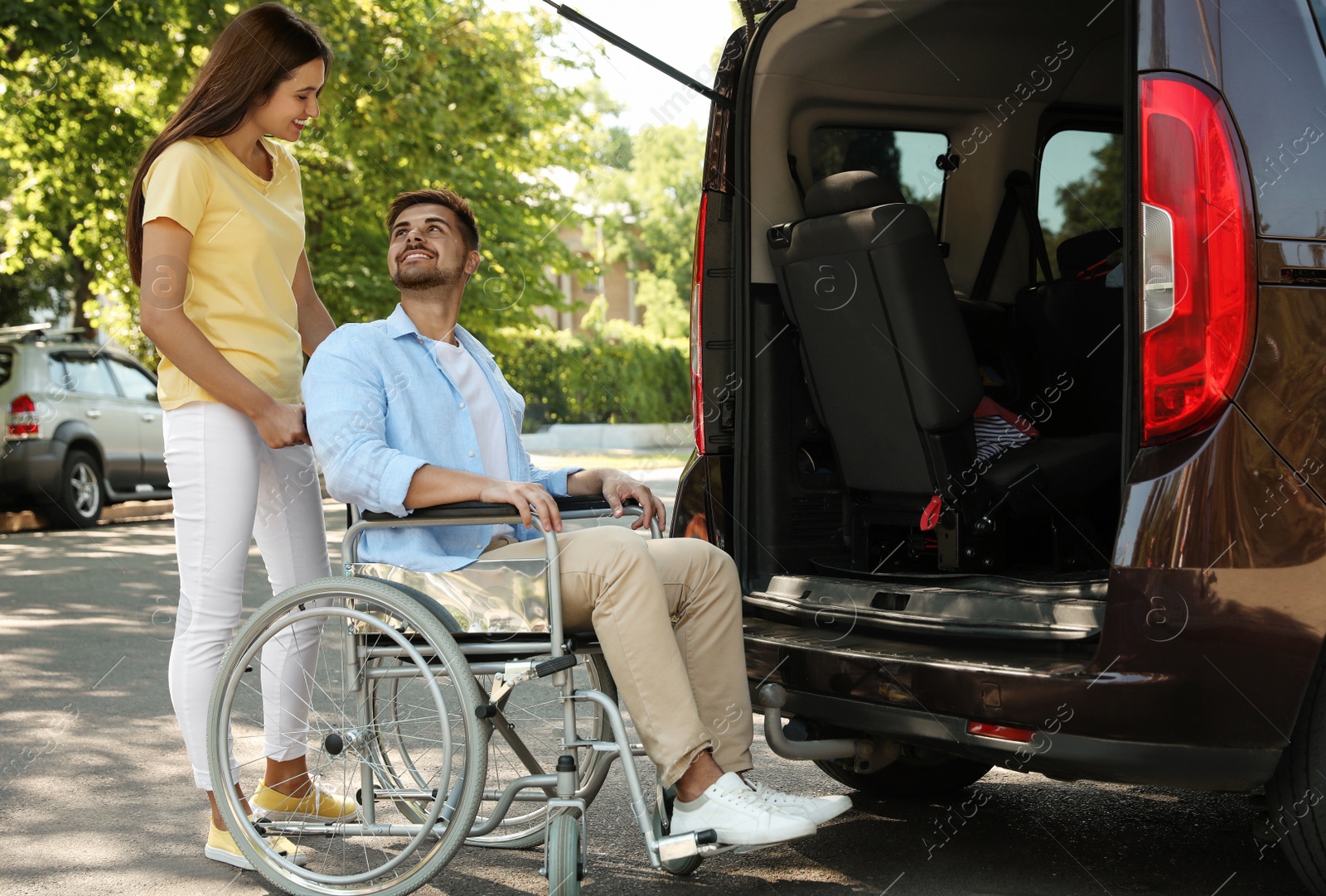 Photo of Young woman helping man in wheelchair to get into van outdoors
