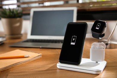 Photo of Mobile phone, earphones and smartwatch charging with wireless pad on wooden desk, space for text. Modern workplace device