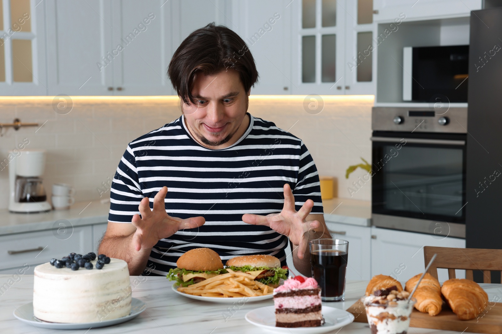 Photo of Hungry overweight man at table with sweets and fast food in kitchen