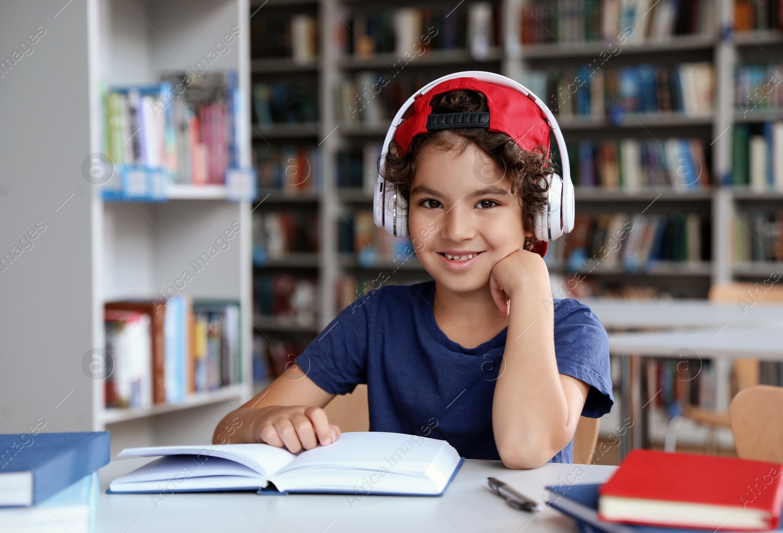 Photo of Cute little boy with headphones reading books at table in library