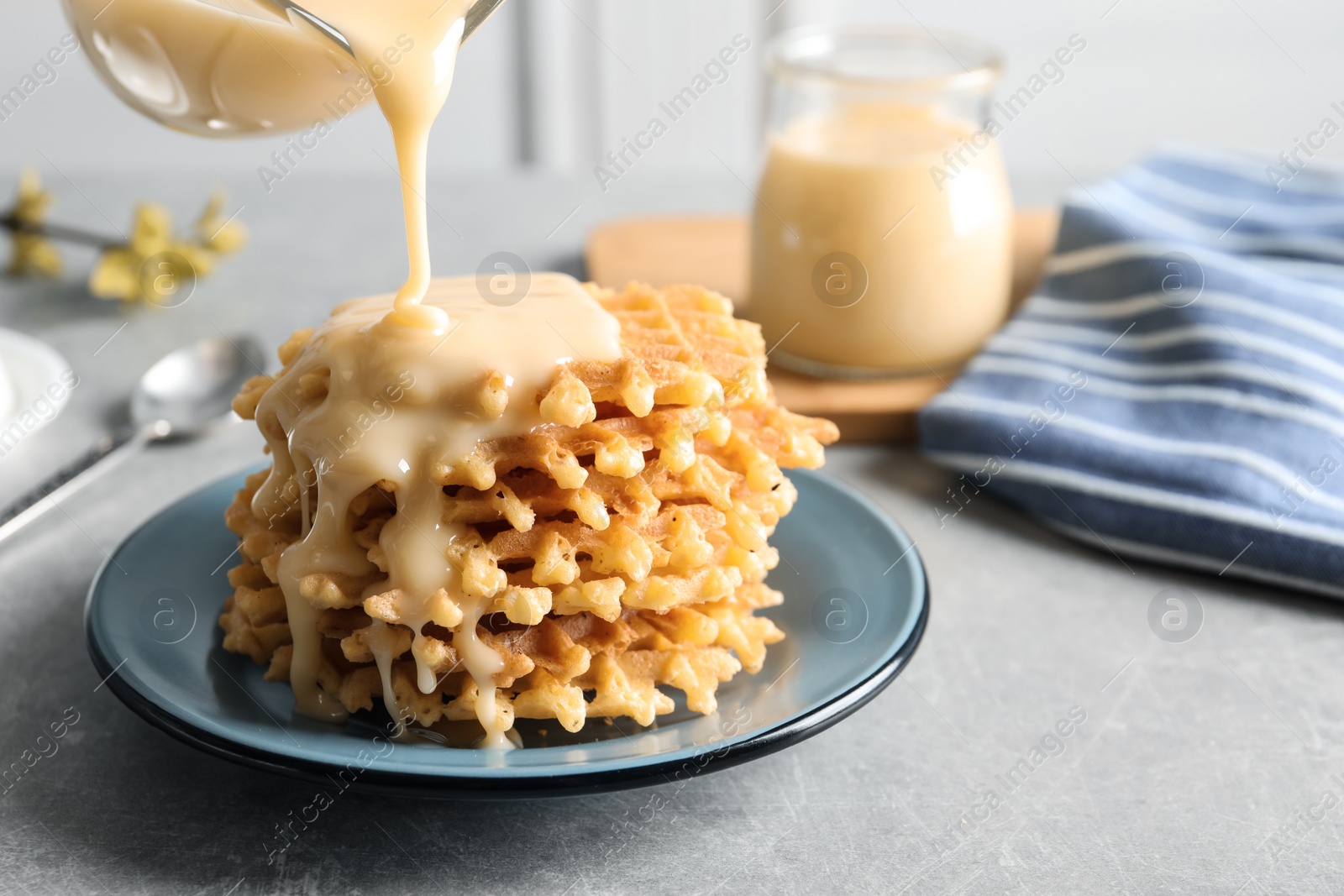 Photo of Pouring condensed milk from jug onto waffles on grey table, space for text. Dairy product