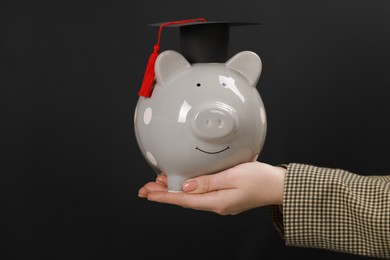 Woman holding piggy bank and graduation cap against black background, closeup. Scholarship concept
