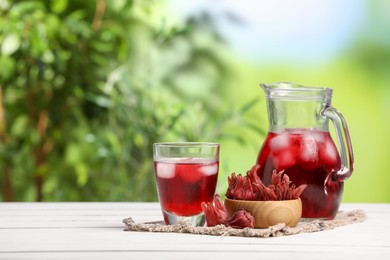Photo of Refreshing hibiscus tea with ice cubes and roselle flowers on white wooden table against blurred green background. Space for text