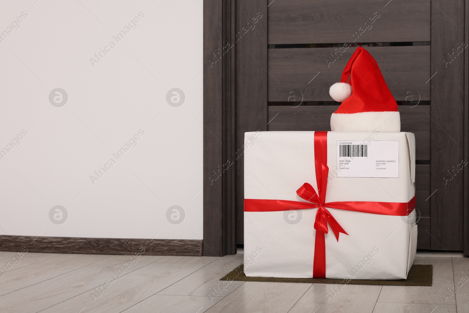 Photo of Christmas gift box and Santa hat near door indoors, space for text. Sending present by mail