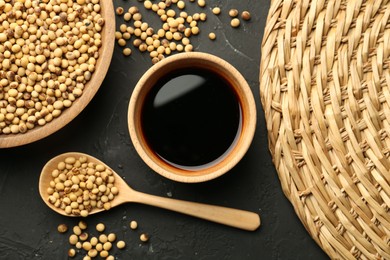 Photo of Soy sauce in bowl and soybeans on black textured table, flat lay