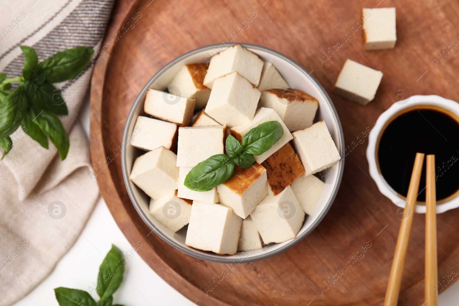 Photo of Bowl of smoked tofu cubes, soy sauce and basil on wooden tray, flat lay