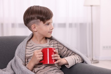 Cute boy under plaid with red ceramic mug on sofa at home, space for text