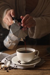 Photo of Woman pouring aromatic coffee from moka pot into cup at wooden table, closeup
