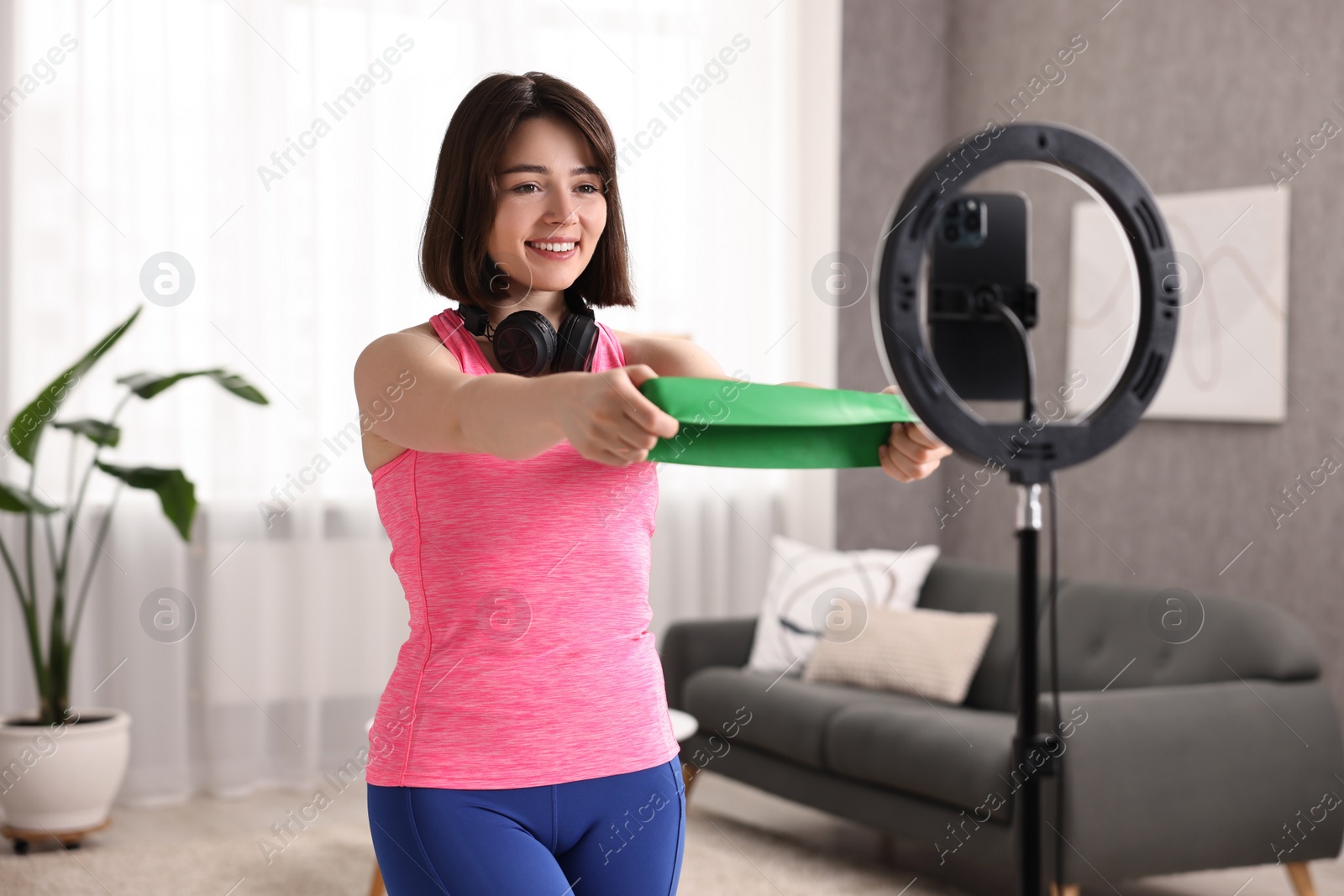 Photo of Happy sports blogger holding resistance band while streaming online fitness lesson with smartphone at home