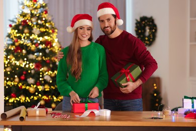 Photo of Happy couple in Santa hats making Christmas gifts at table in room