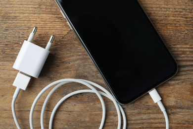 Smartphone and charging cable with adapter on wooden table, flat lay