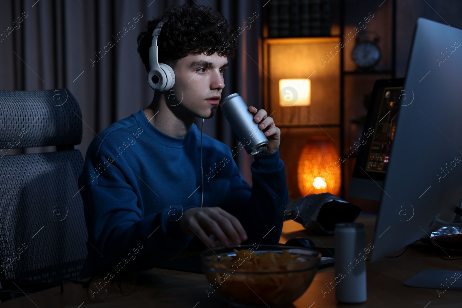 Photo of Young man with energy drink and headphones playing video game at wooden desk indoors