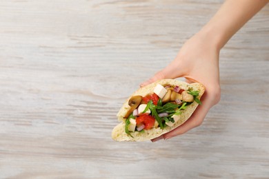 Woman holding delicious pita sandwich with grilled vegetables and parsley at light wooden table, top view. Space for text