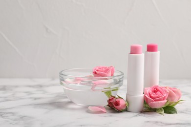 Photo of Lip balms, bowl with water and roses on white marble table, space for text