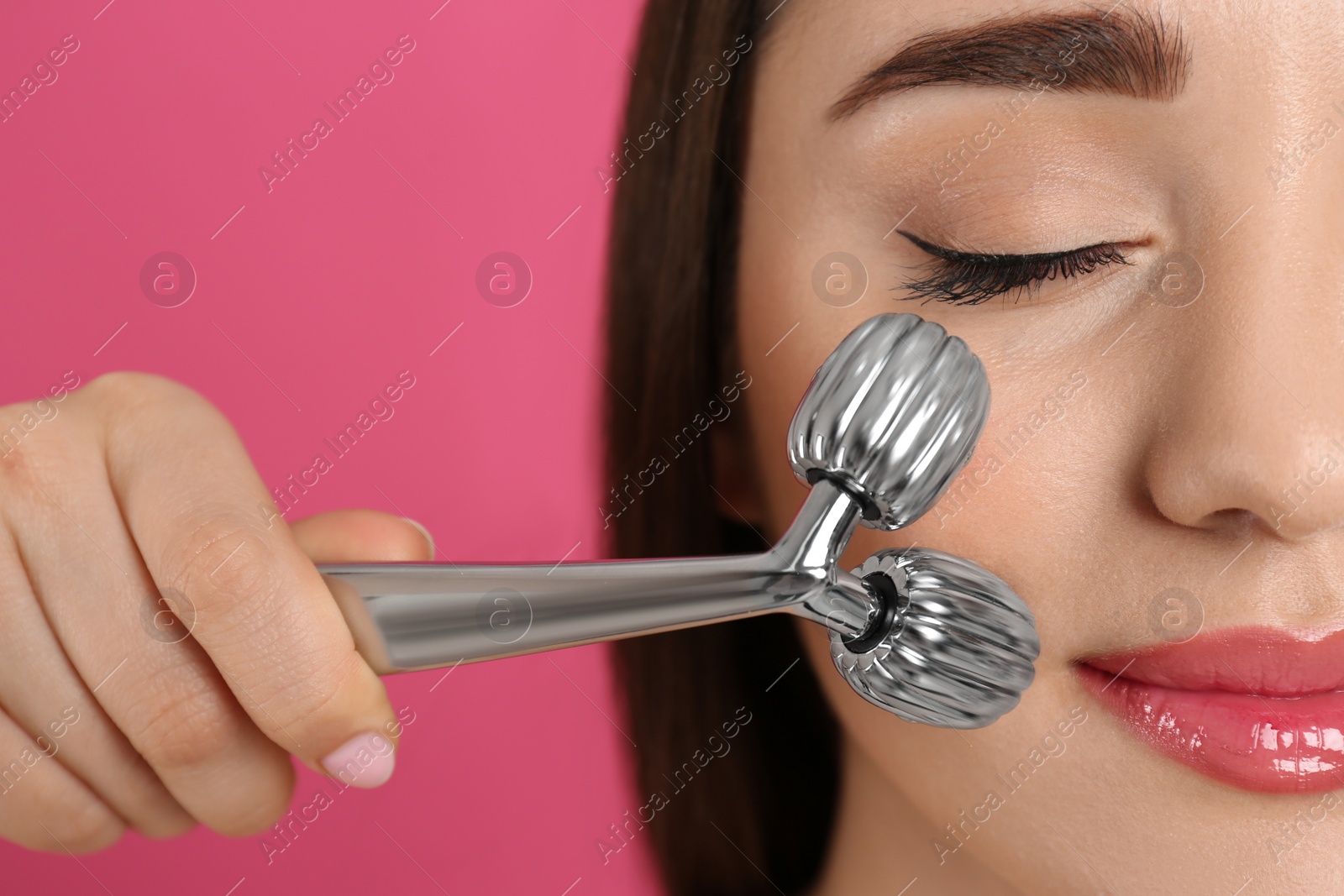 Photo of Woman using metal face roller on pink background, closeup
