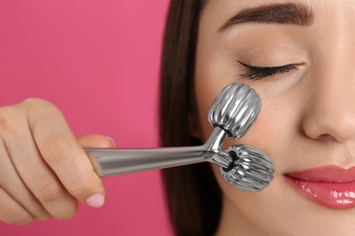 Woman using metal face roller on pink background, closeup