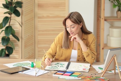 Young woman painting green twig in sketchbook at wooden table indoors