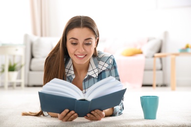 Beautiful young woman with cup of coffee reading book on floor at home