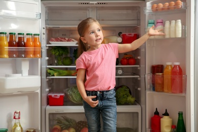 Photo of Cute little girl standing near open refrigerator at home