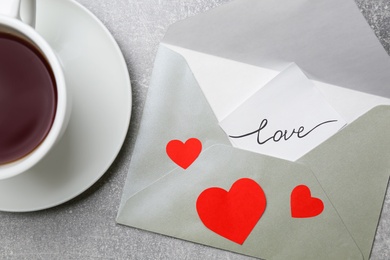Photo of Card with handwritten word love in envelope near cup of coffee on grey table, flat lay