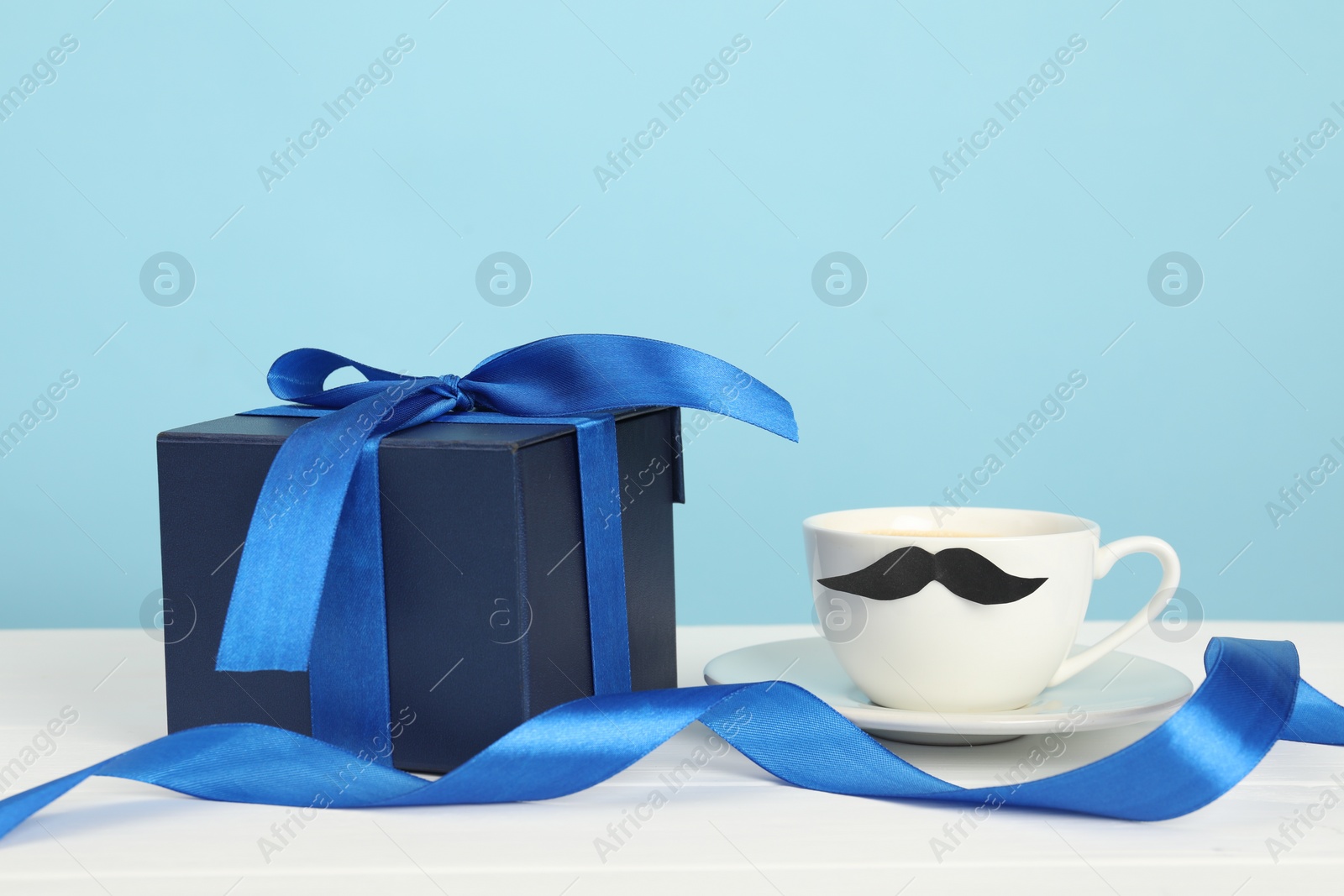 Photo of Happy Father's Day. Cup with moustache and gift box on white wooden table, closeup