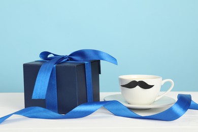 Happy Father's Day. Cup with moustache and gift box on white wooden table, closeup