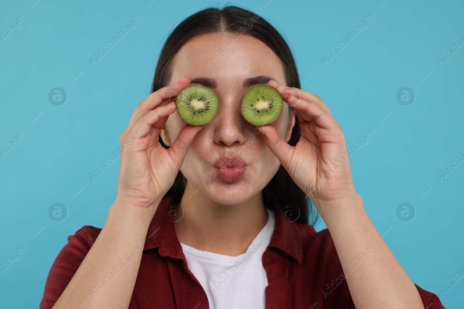 Photo of Woman covering eyes with halves of kiwi on light blue background