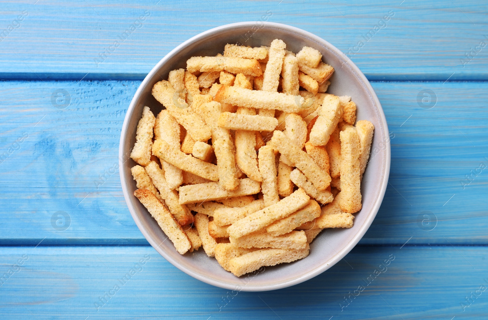 Photo of Crispy wheat rusks in bowl on light blue wooden table, top view