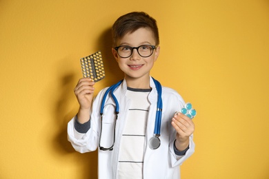 Cute child in doctor coat with pills on color background