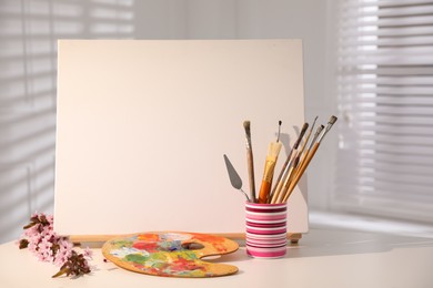 Photo of Easel with blank canvas, fresh flowers and art supplies on white table