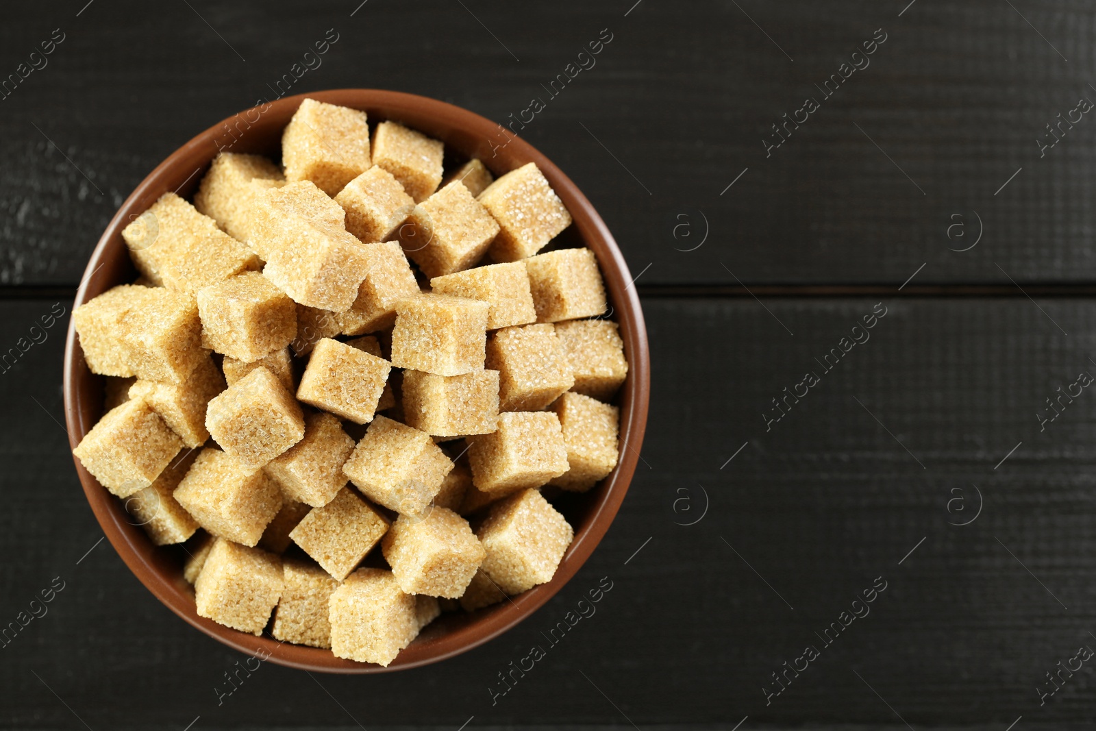 Photo of Brown sugar cubes in bowl on black wooden table, top view. Space for text