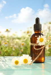 Photo of Bottle of chamomile essential oil on light blue wooden table in field