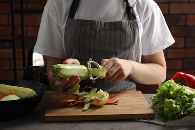 Photo of Woman peeling fresh zucchini at grey table indoors, closeup