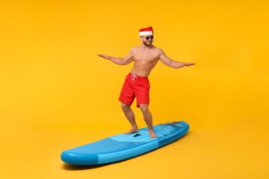 Happy man in Santa hat balancing on SUP board against orange background