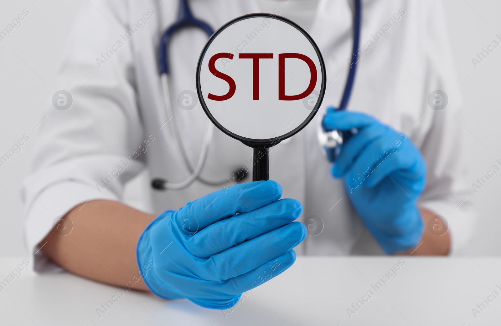 Photo of Doctor holding magnifying glass with abbreviation STD at white table, closeup