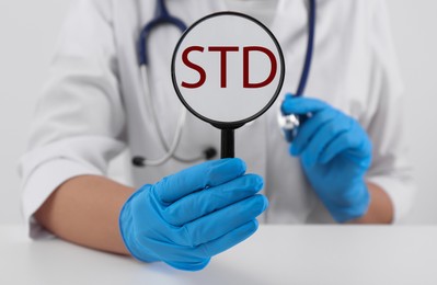 Photo of Doctor holding magnifying glass with abbreviation STD at white table, closeup