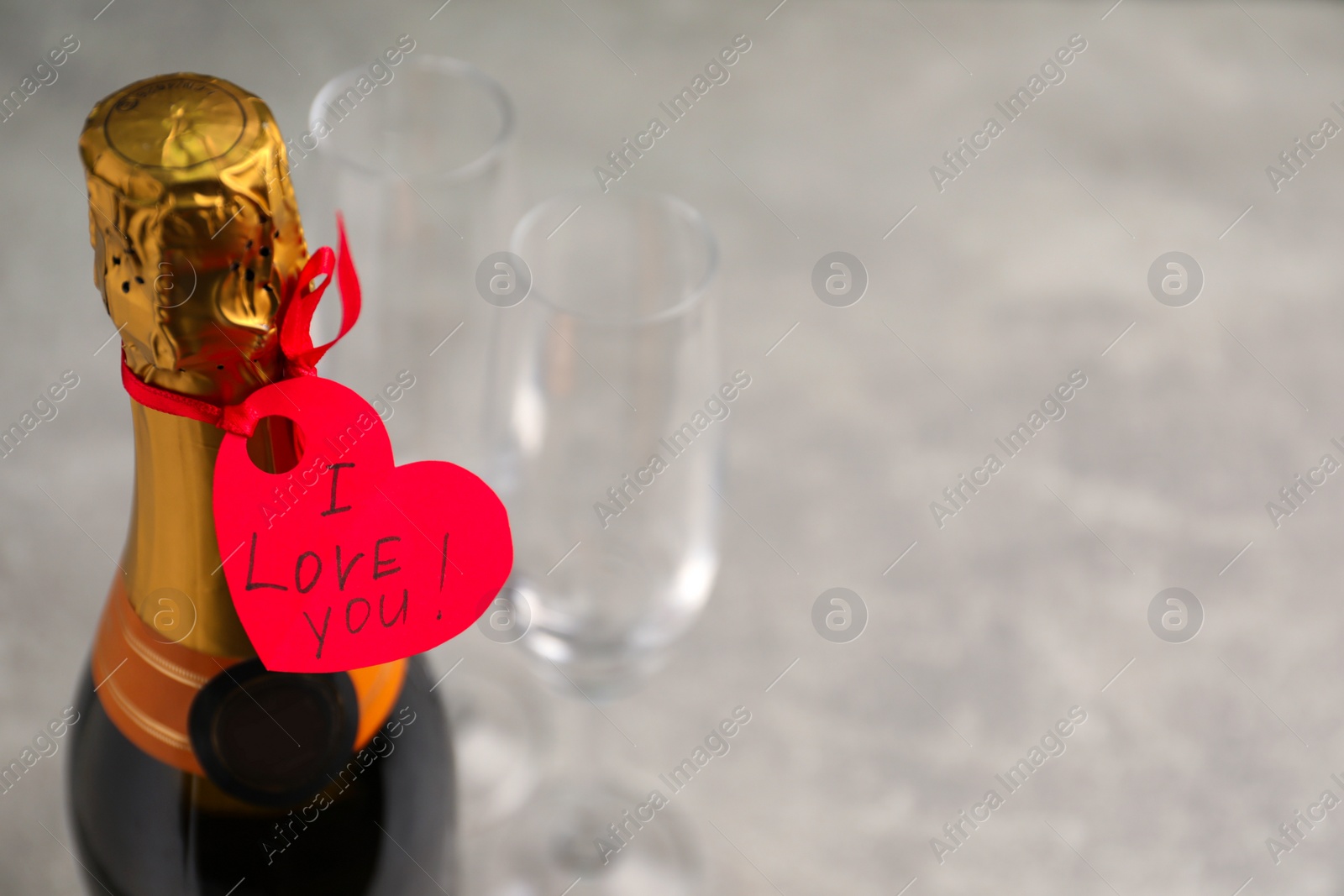 Photo of Heart shaped greeting card with phrase I Love You tied to bottle of champagne on light table, closeup. Space for text