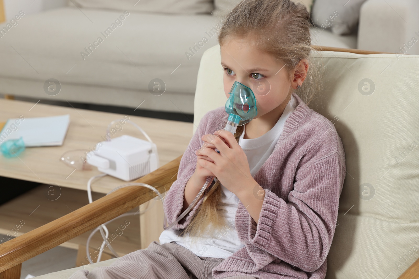 Photo of Little girl using nebulizer for inhalation in armchair at home