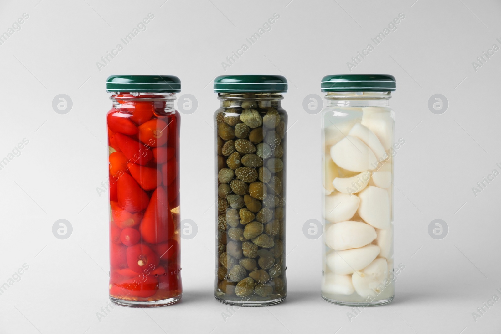 Photo of Jars of pickled red hot piri-piri peppers, capers and garlic on light grey background