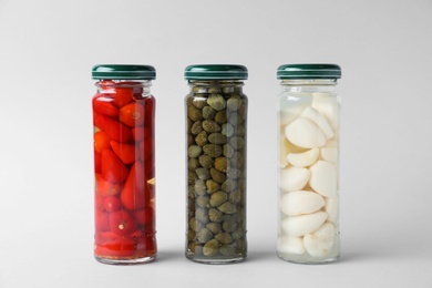 Jars of pickled red hot piri-piri peppers, capers and garlic on light grey background