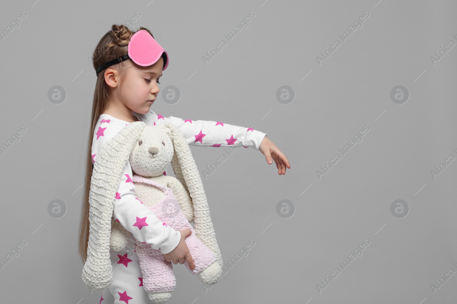 Photo of Girl in pajamas and sleep mask with toy bunny sleepwalking on gray background, space for text