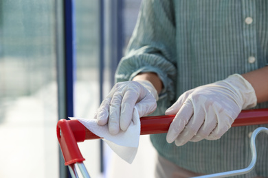 Woman cleaning handle of shopping cart with wet wipe on blurred background, closeup